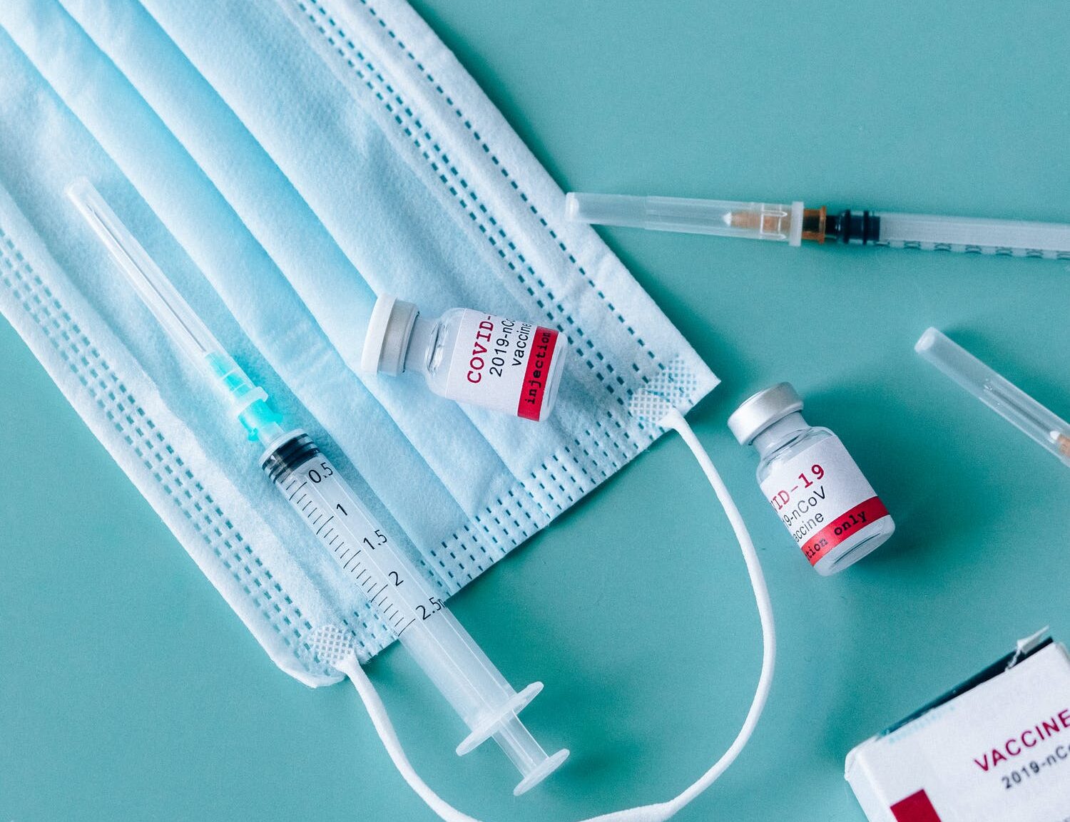 Munskydd, vaccin och sprutor som ligger på ett turkost bord