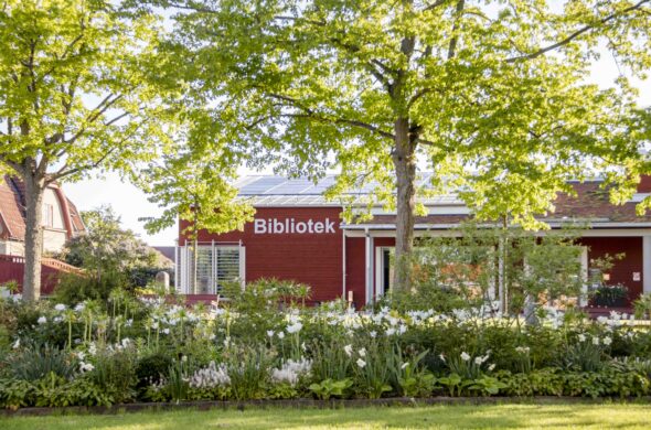 Grönskade blomsterrabatter och träd. Bakom träden syns en röd byggnad med texten Bibliotek på fasaden.