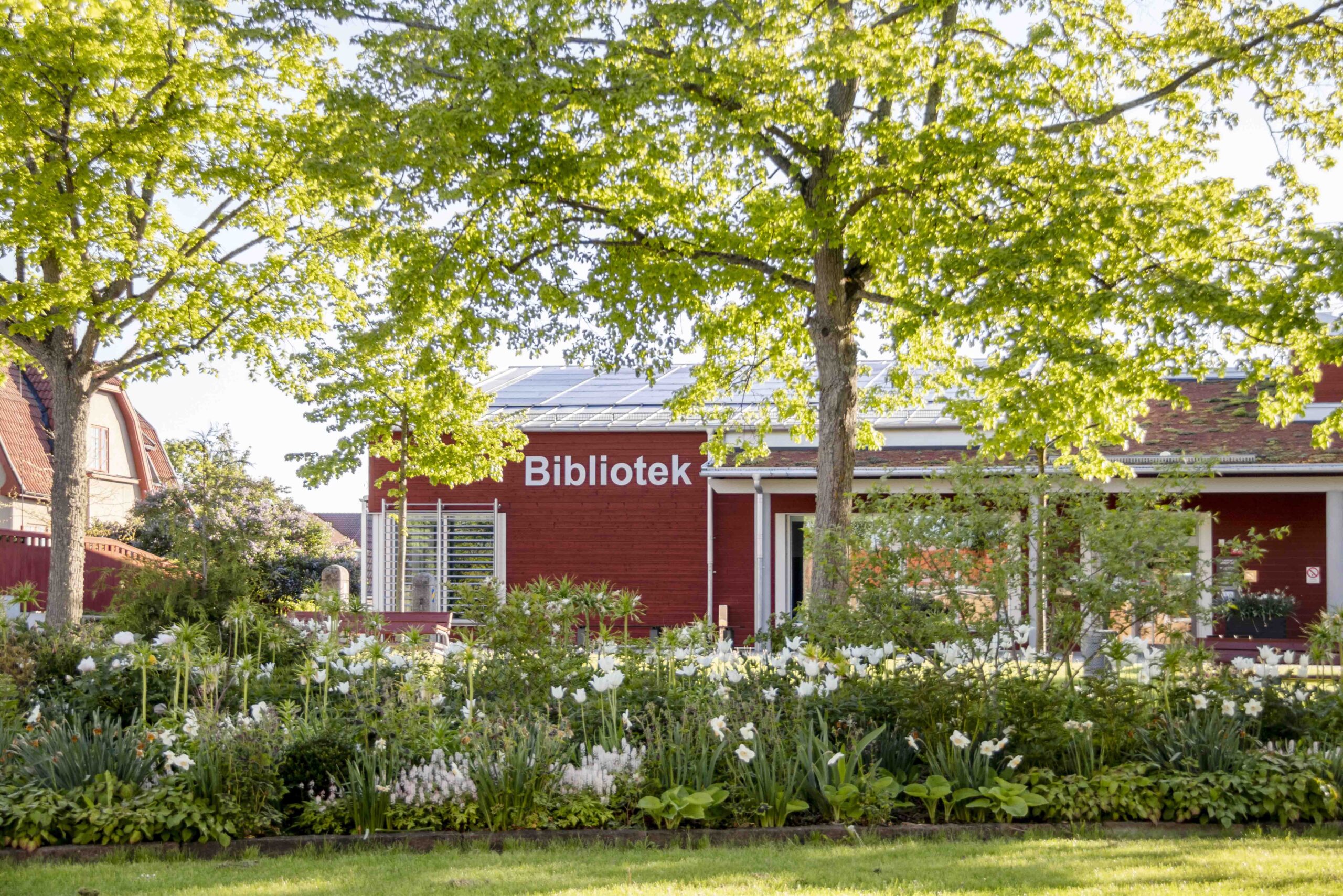 Grönskade blomsterrabatter och träd. Bakom träden syns en röd byggnad med texten Bibliotek på fasaden.