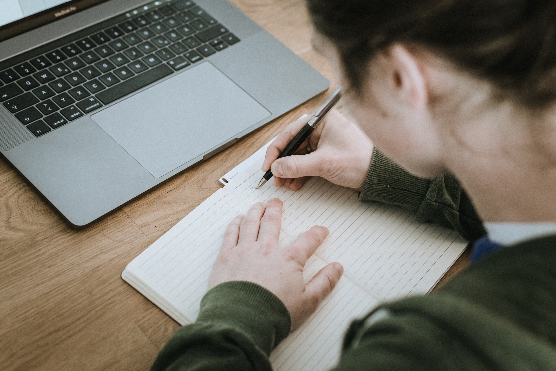 En person skriver i en anteckningsbok med en dator framför sig