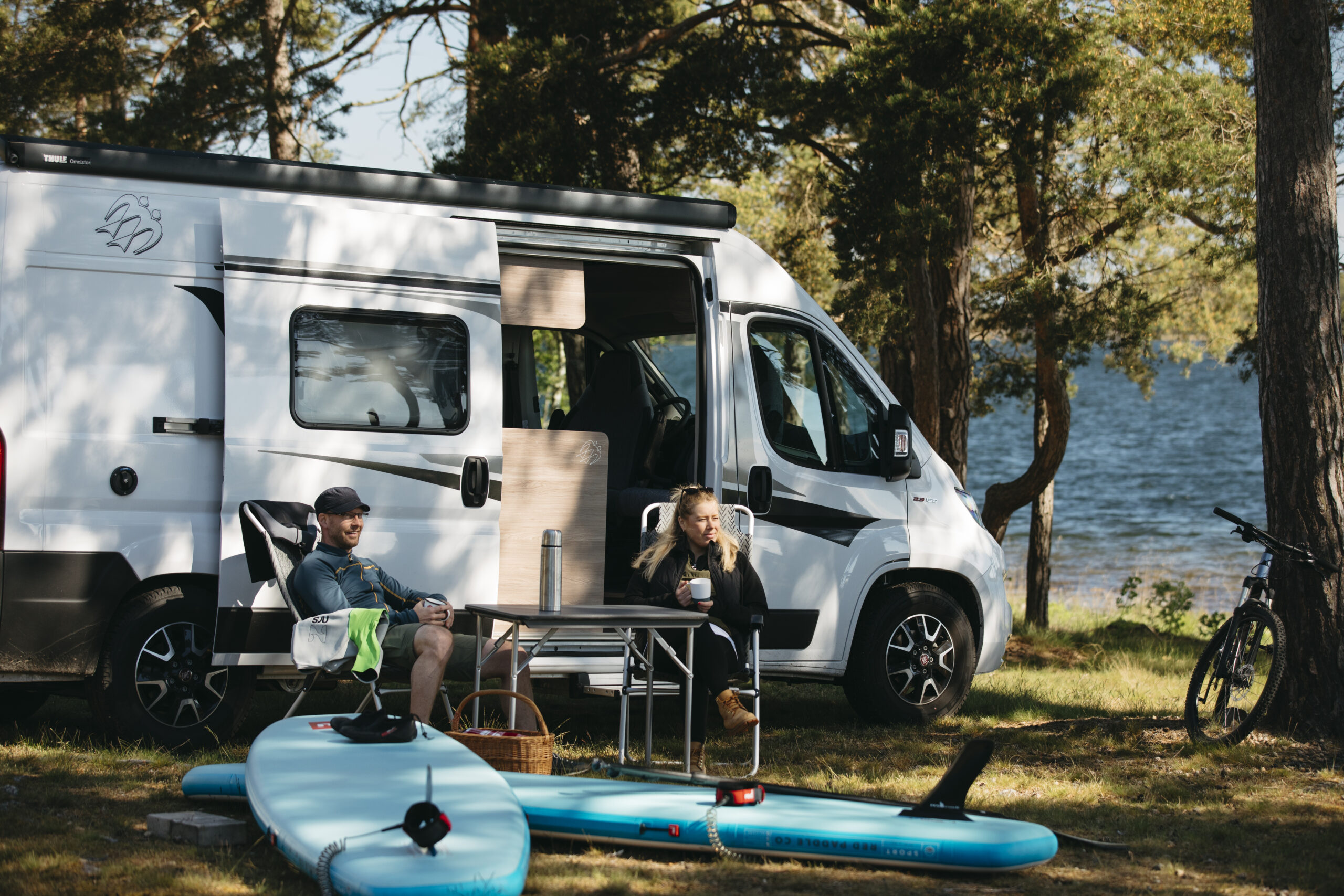 Två personer sitter i campingstolar framför paddle boards, i bakgrunden syns en husbil och hav