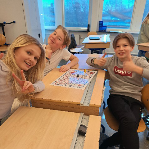 Elever sitter vid några bänkar i deras klassrum och spelar ett brädspel. Alla är glada och ler in mot kameran.