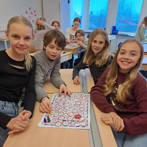 Elever sitter vid några bänkar i deras klassrum och spelar ett brädspel. Alla är glada och ler mot kameran. I bakgrunden syns andra elever som även de spelar.