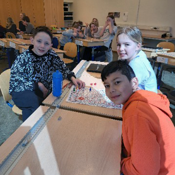 Elever sitter vid några bänkar i deras klassrum och spelar ett brädspel. Eleverna är glada och ler in mot kameran. I bakgrunden syns andra grupper med elever som även de spelar.
