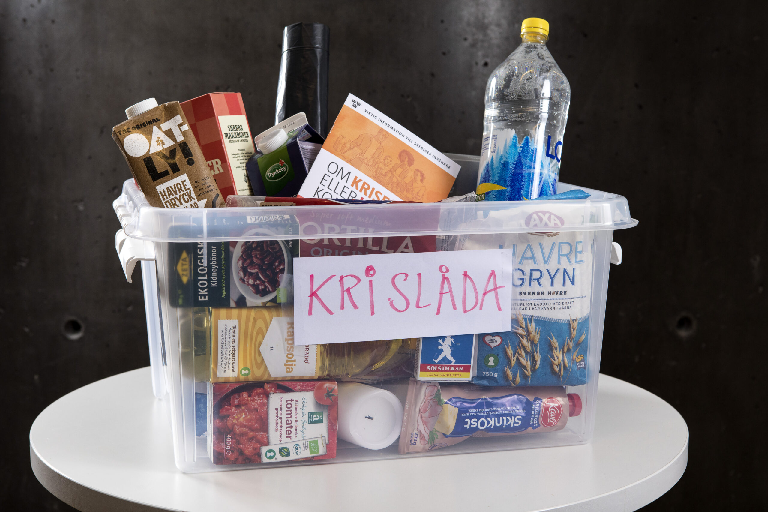 En genomskinlig plastlåda står på ett podium. På en lapp på utsidan av lådan står ordet Krislåda. Lådan är fylld med bra saker att ha hemma vid en kris, till exempel vatten, stearinljus och olika matvaror.