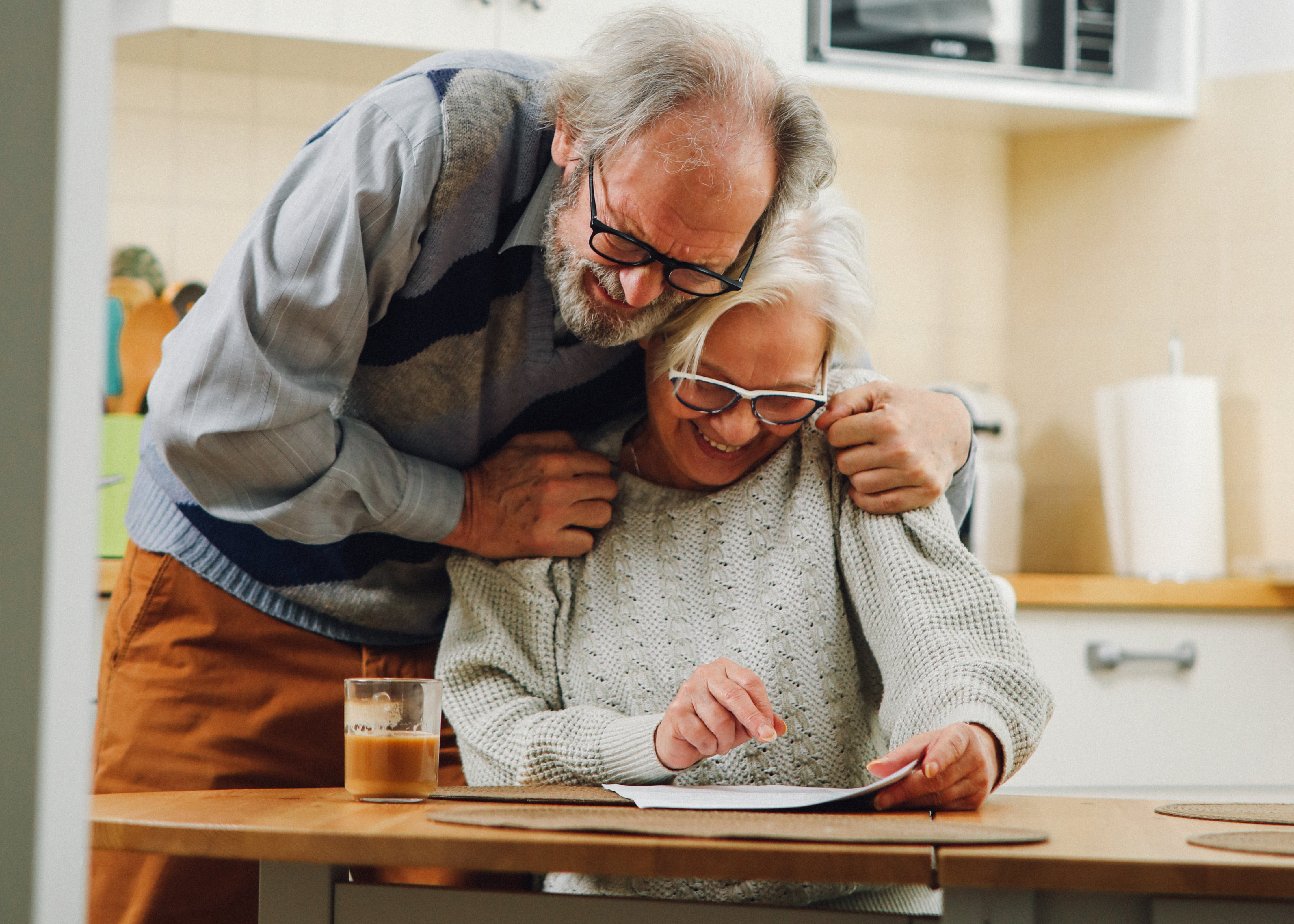 Foto. Ett äldre par med en kvinna som sitter vid ett köksbord och en man som står bakom och kramar om henne. Båda tittar ner på bordet och ler, i handen håller kvinnan ett papper,