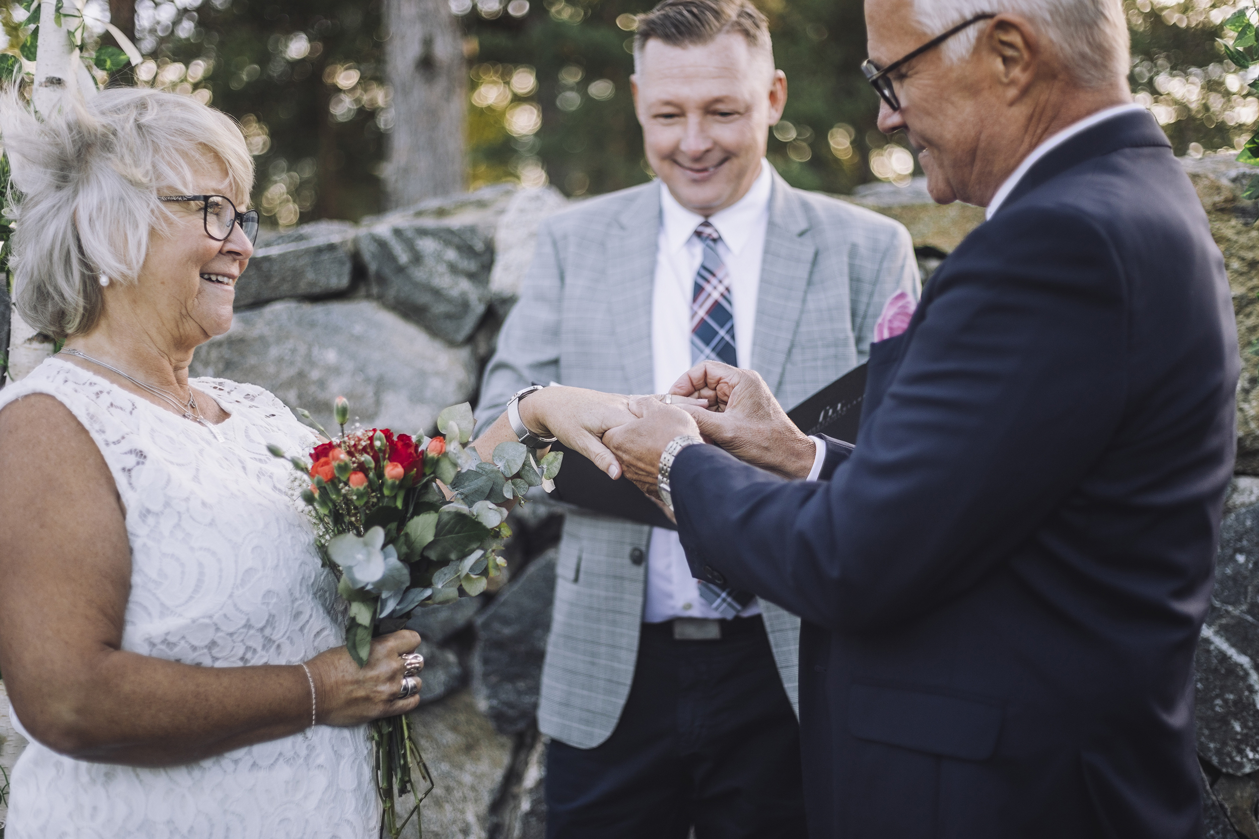 Äldre par gifter sig på en äng. Mannen trär på bruden ringen. I bakgrunden står vigselförrättaren.