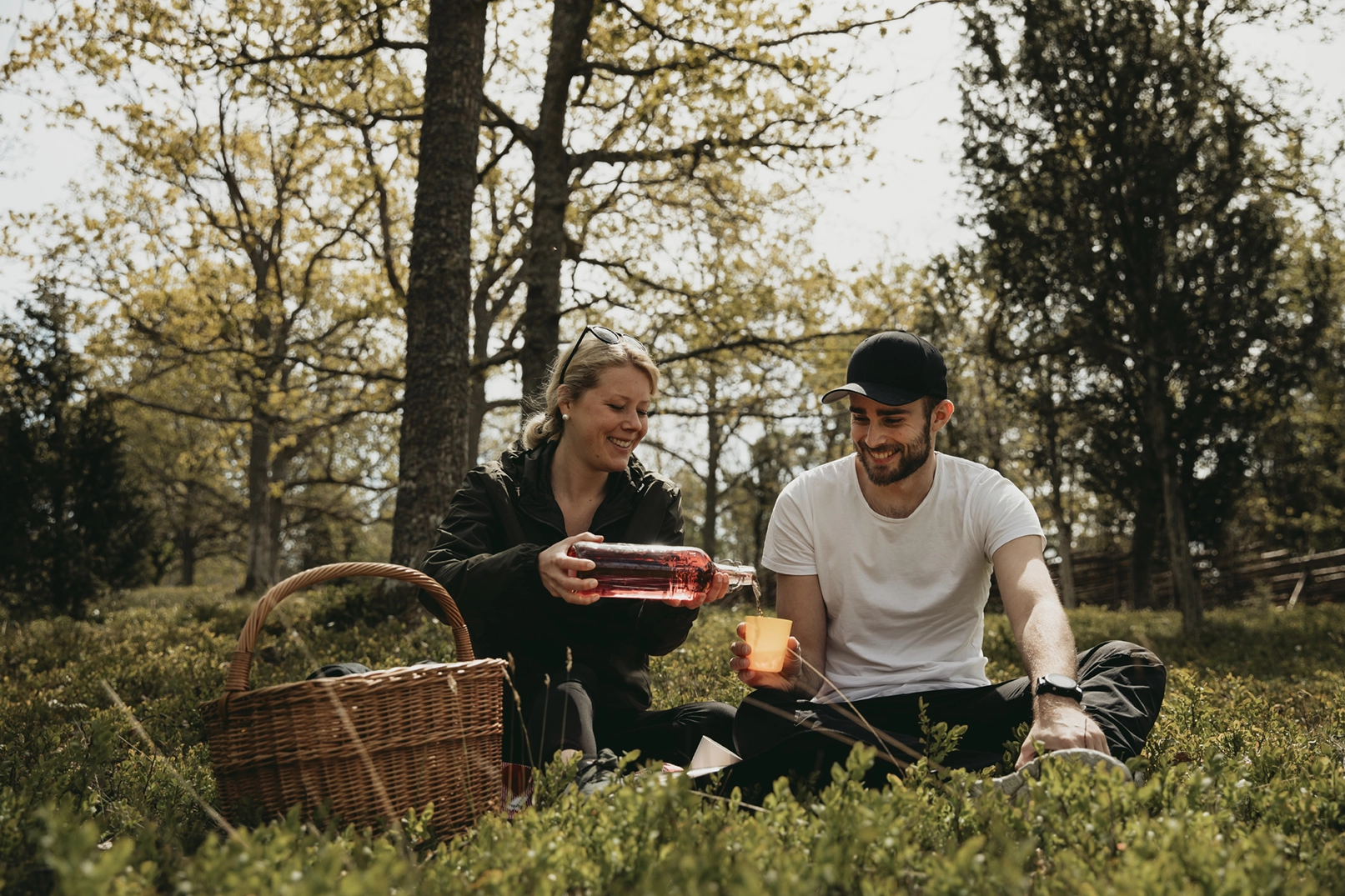 En man och en kvinna sitter på en filt i gröngräset och har picknick. Kvinnan häller upp saft i ett glas som mannen håller fram.