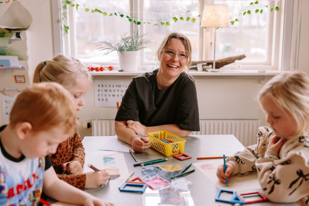 Leende förskollärare sitter vid ett bord med tre barn som ritar
